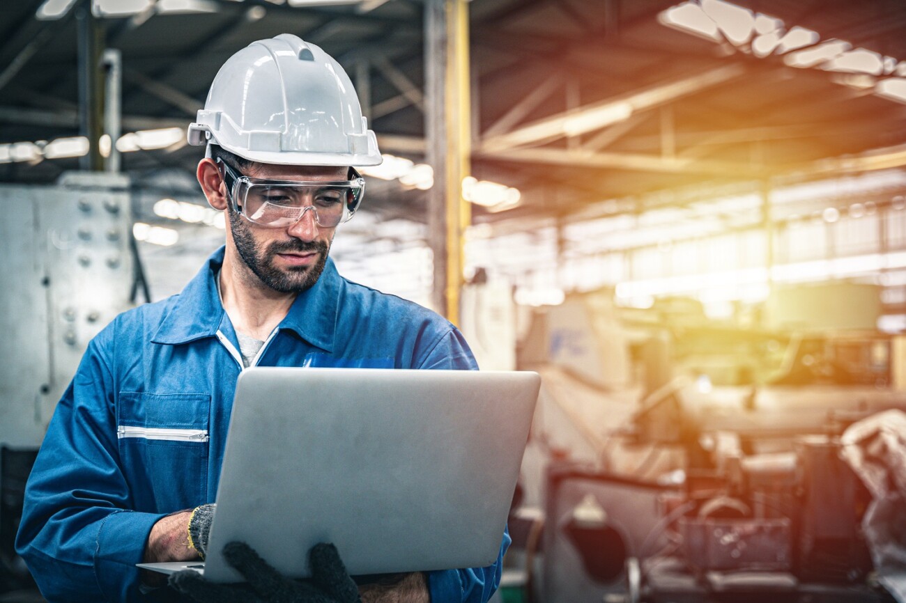 Confident Engineer In Blue Jumpsuit Holding Laptop Computer In A Warehouse.