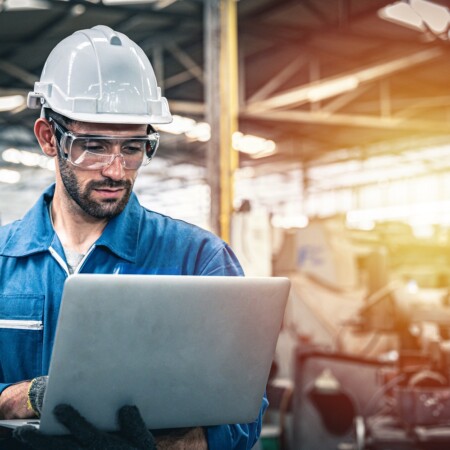 Confident Engineer In Blue Jumpsuit Holding Laptop Computer In A Warehouse.